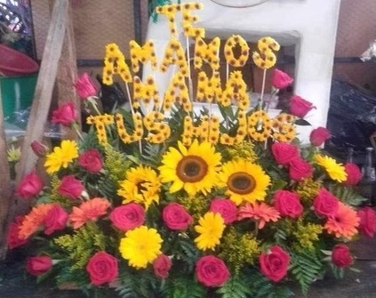 An image of pink and yellow flowers in an arangement with yellow sunflowers arranged to spell out Te Amos Mama tus hijos