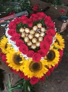 An image of red roses and yellow sunflowers made into the shape of a heart with gold chocolates in the middle